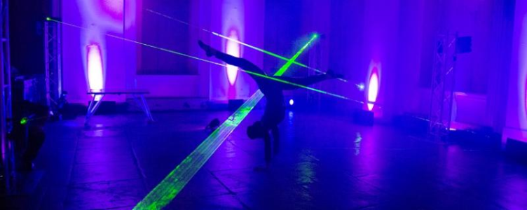 Acrobat navigating through a series of green laser beam lights in a room with purple uplighting
